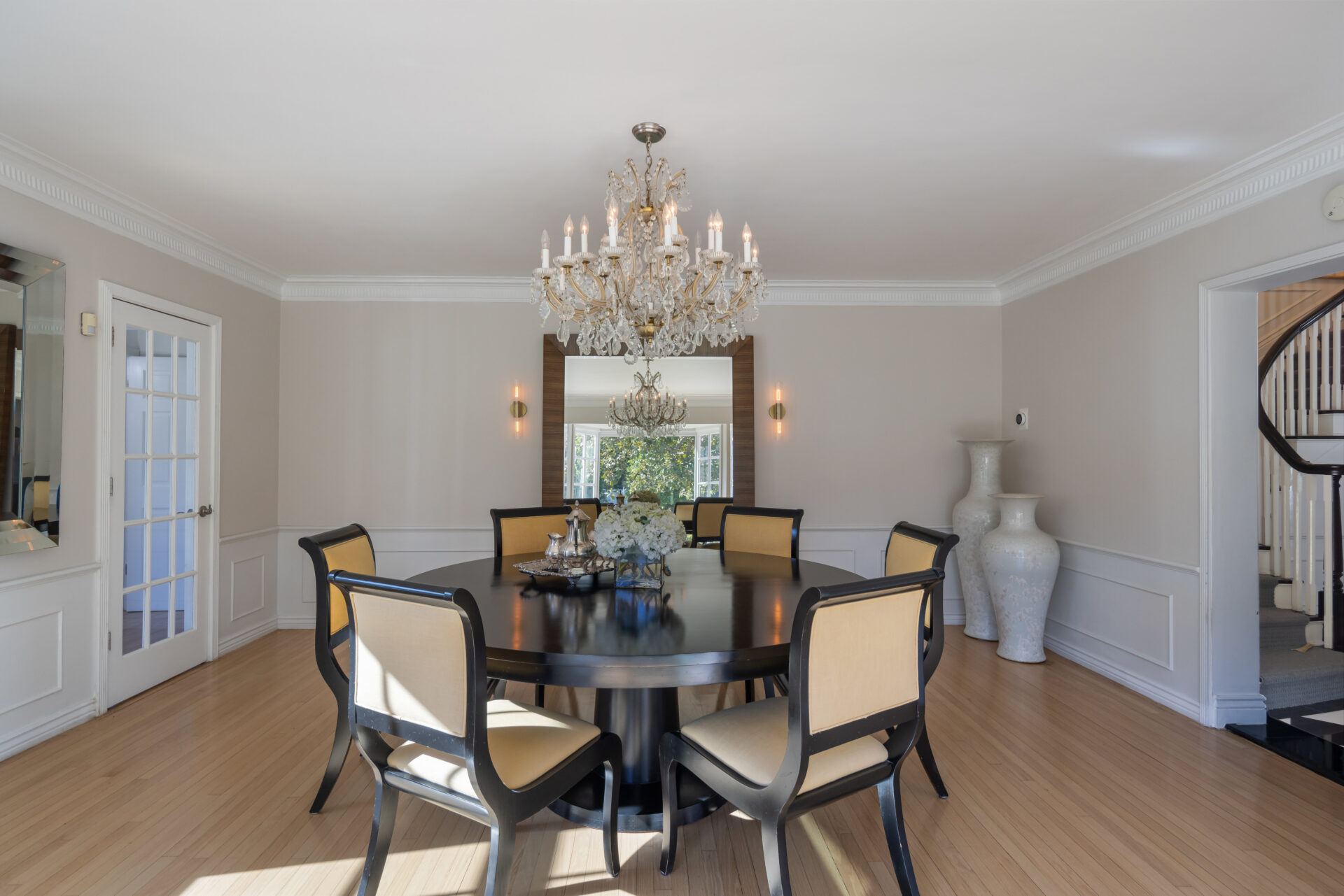 A large crystal chandelier hangs above a round dining table, while a wide mirror along the far wall reflects light from the windows and adds depth to the room.
