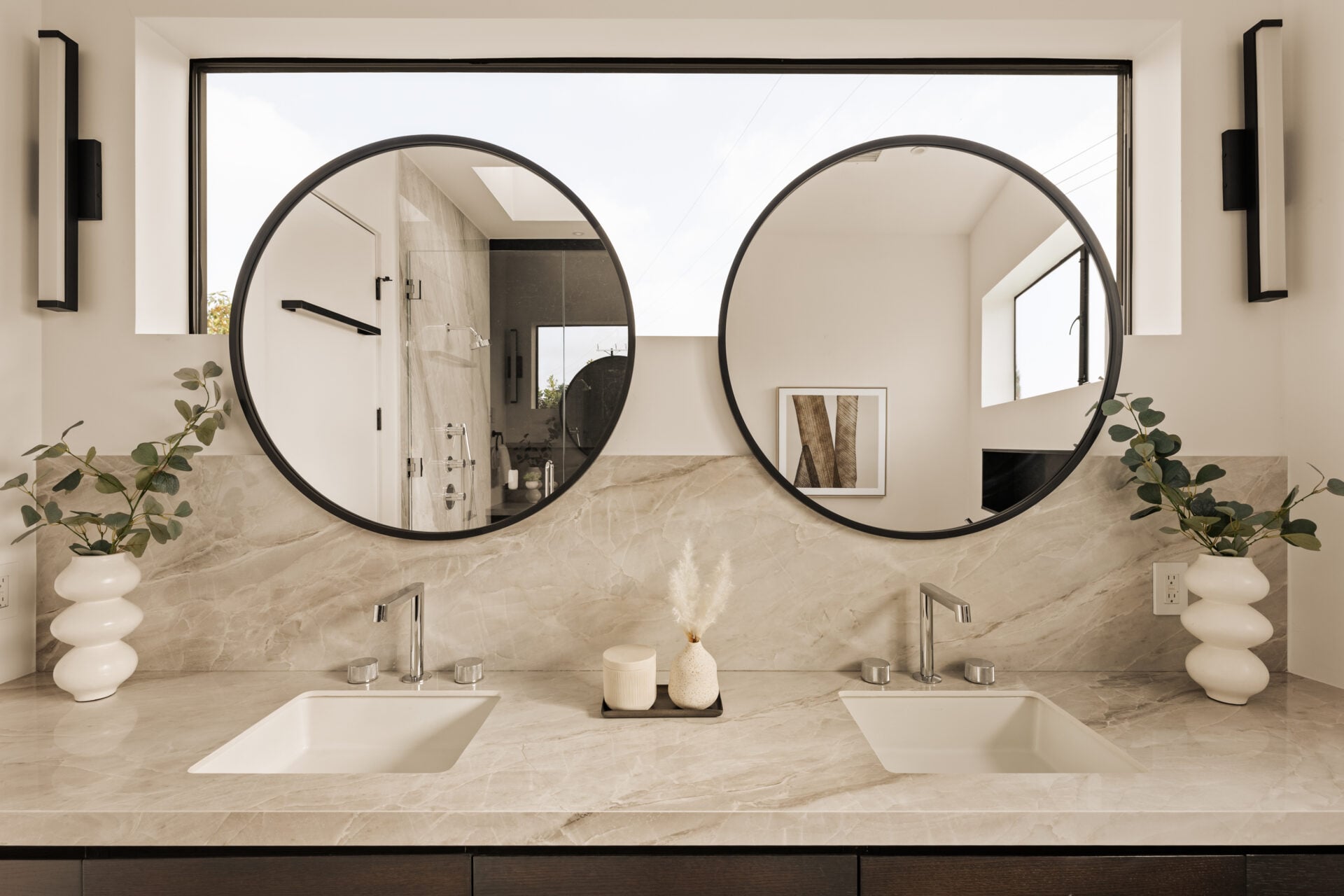 A long double vanity finished in pale stone, with two sinks set beneath oversized round mirrors and a horizontal window.