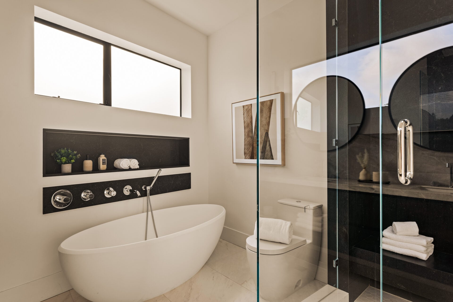 A freestanding soaking tub sits beneath a horizontal window, with a recessed shelf built into the wall above it for storage and display.