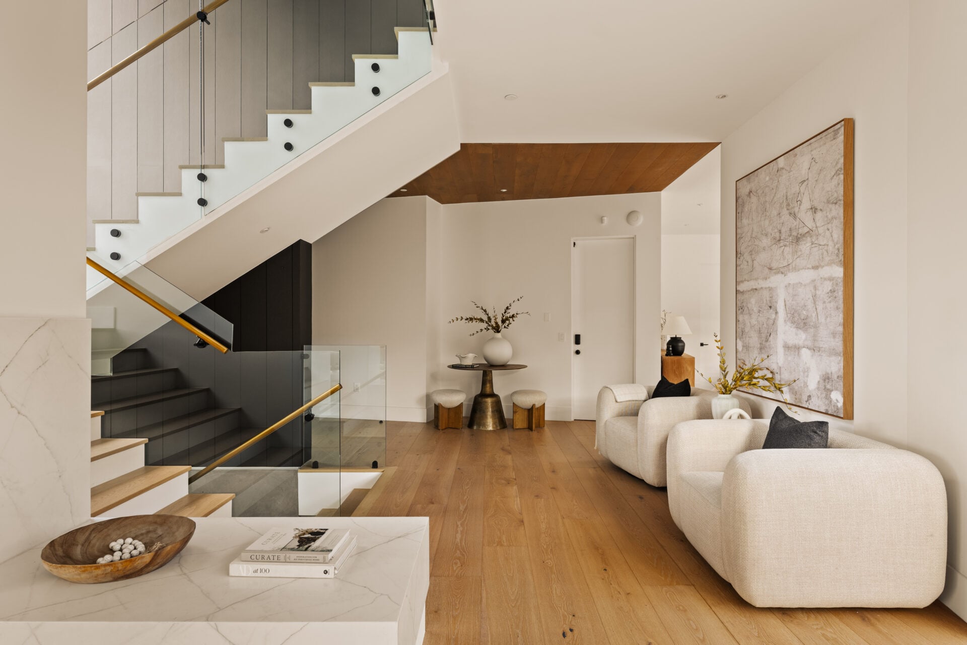 A glass-railed staircase rises along one side while a pair of low upholstered chairs sit across from it beneath oversized artwork.