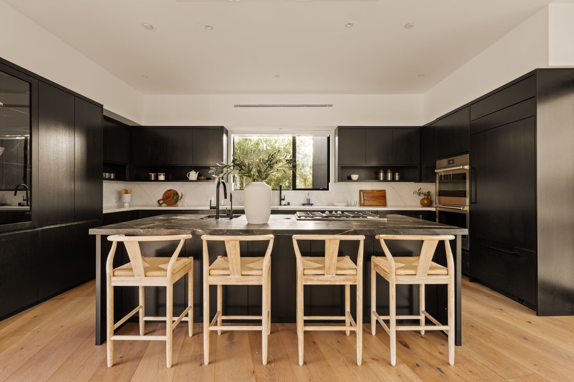 At the center, an oversized natural quartzite island has a sink and the stovetop on its inner side four chairs placed along the outer edge.