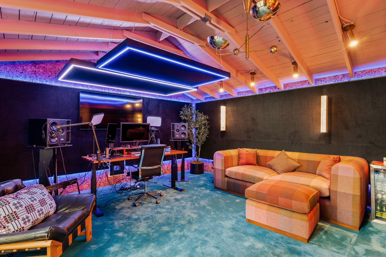 The main recording room is set beneath exposed beams with acoustic treatments lining the walls around multiple workstations, monitors, microphones, and a large screen.