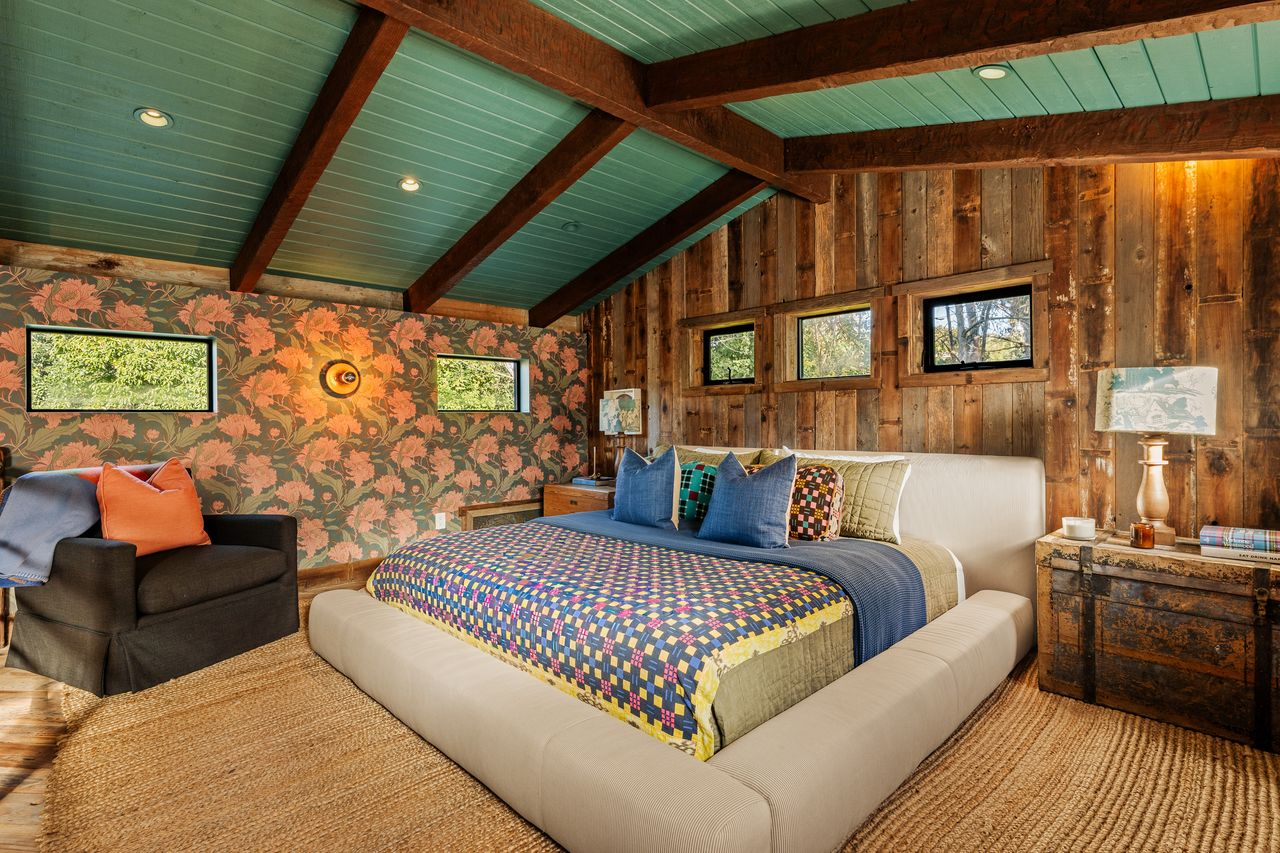 Exposed beams and wood continue overhead and along the walls, while a row of smaller windows brings in treetop views around the bed.