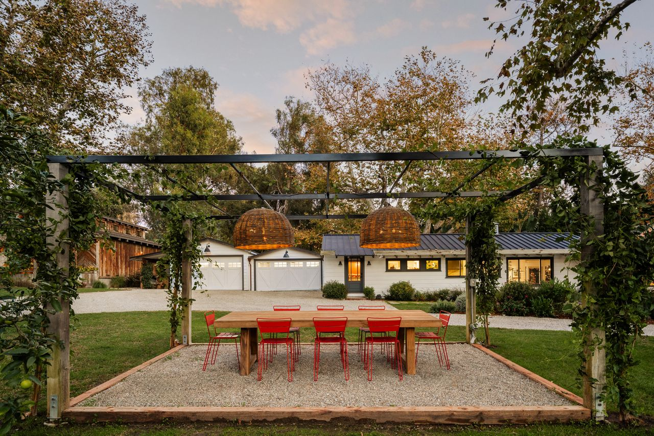 Across the driveway, a long outdoor dining table sits beneath a vine-covered pergola, creating a separate place for meals away from the house.