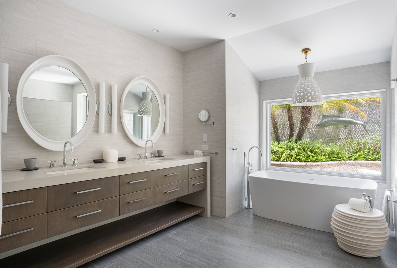 Dual vanities next to a freestanding soaking tub sits beneath a wall-length window overlooking the tropical landscaping.