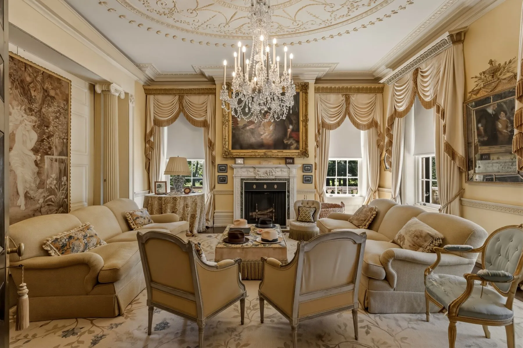 A marble fireplace is centered along the far wall, with a large gold-framed painting rising above it and tall windows with heavy drapery on either side. 