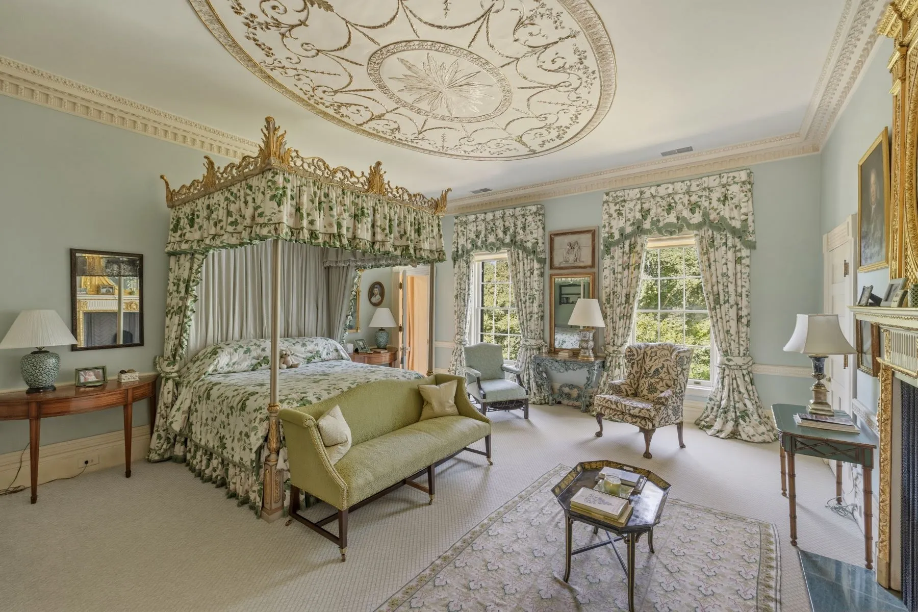 Bedroom with a softer green palette, with floral fabrics lining the canopy bed, window curtains, and surrounding seating. 