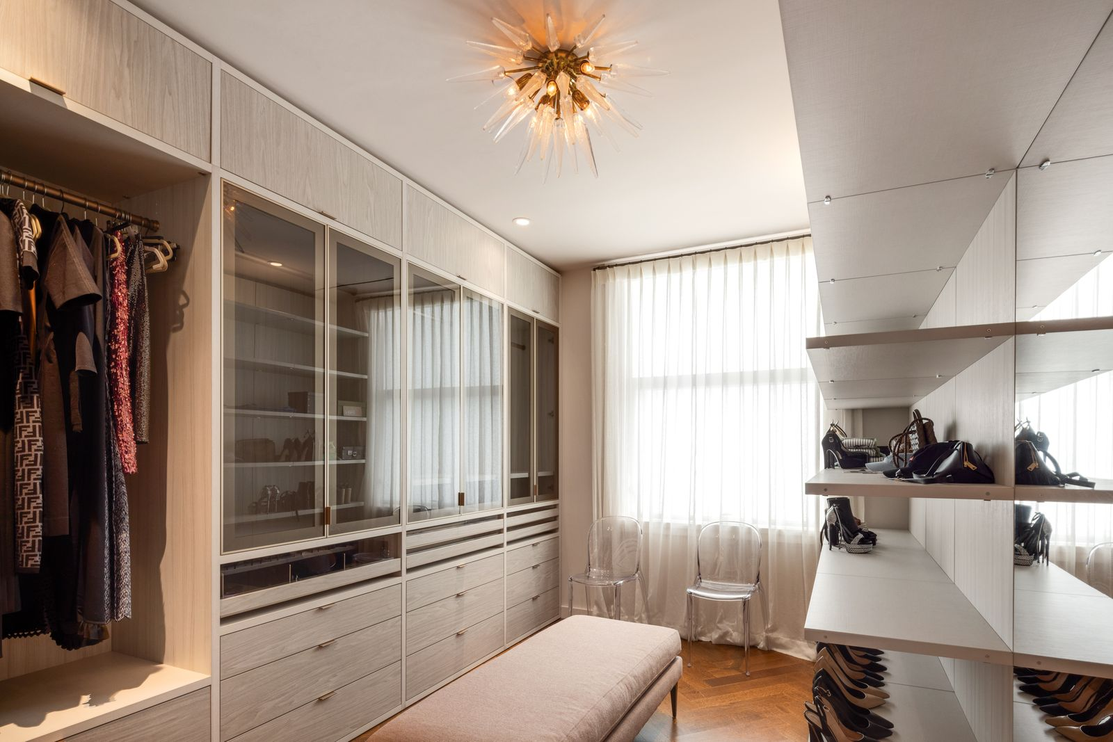 Floor-to-ceiling custom cabinetry lines one wall with a mix of hanging storage, drawers, and glass-front display cases, while mirrored shelving on the other wall adds room for shoes and accessories.