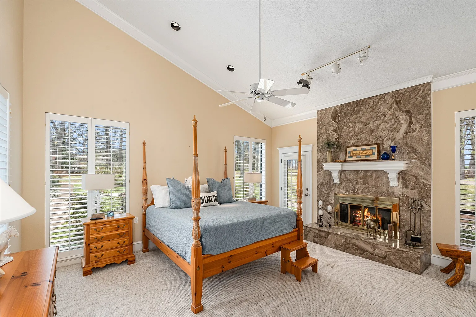 Tall shuttered windows line both sides of the bedroom, bringing in views of the surrounding grounds, while the far wall has a floor-to-ceiling stone fireplace.