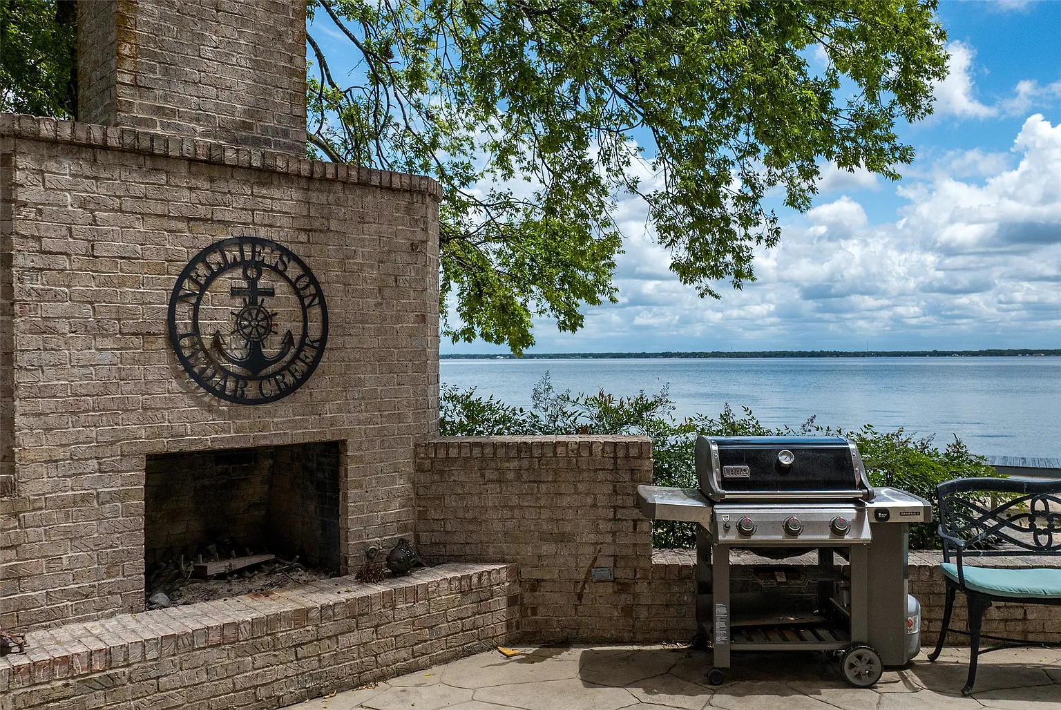 A brick-lined outdoor kitchen sits along the edge of the patio with an open fireplace built into one side and a grill positioned beside it.