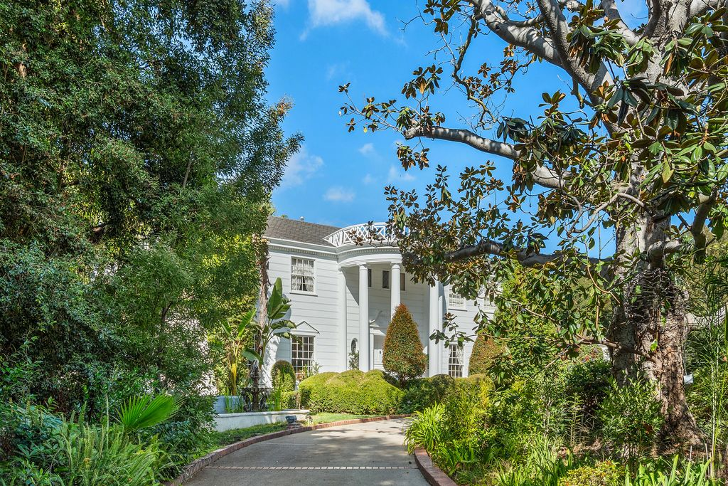 The tall columns frame the main entrance beneath a curved upper balcony, while trimmed hedges and plants line the walkway.