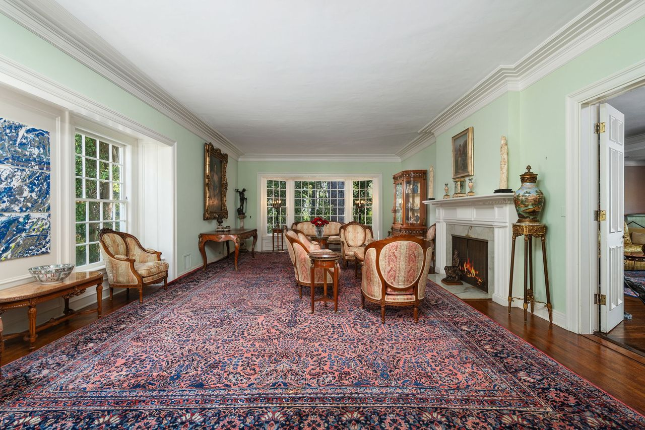 A fireplace sits along the right wall beneath a traditional mantel, with formal couches and chairs spread around the room atop a deep-colored patterned rug.