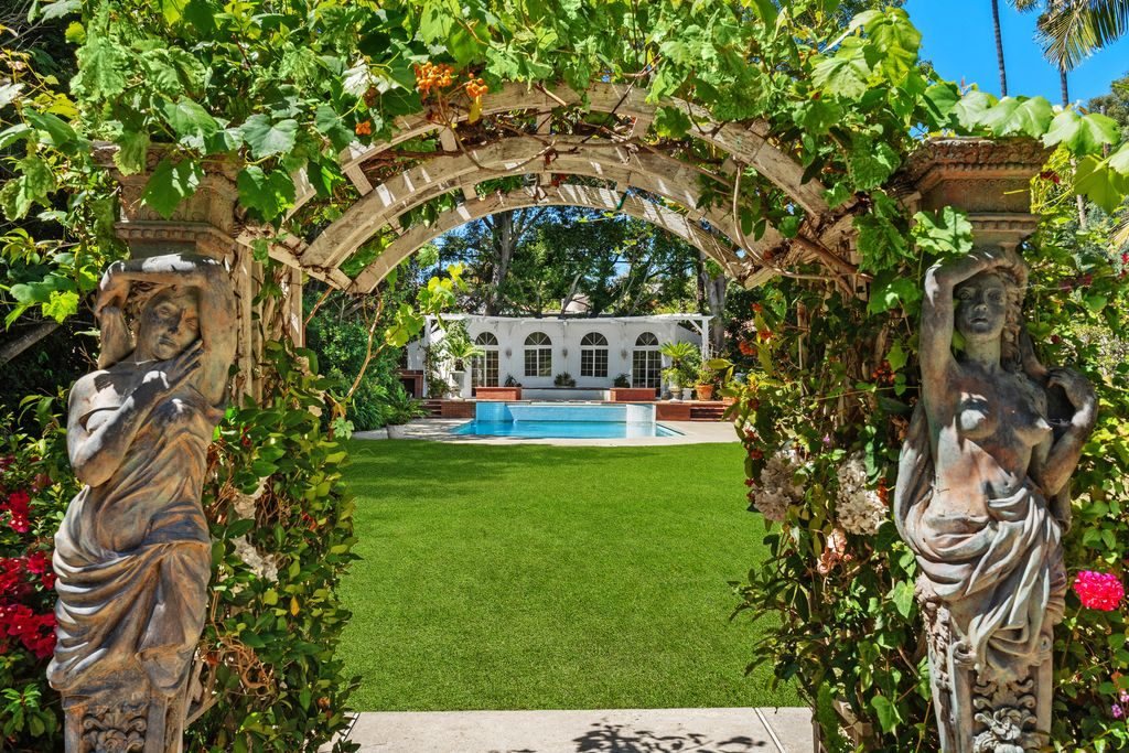 A stone arbor marks the transition from the lower patio into the main lawn, with climbing vines wrapping over its curved frame and sculptural figures set along either side.