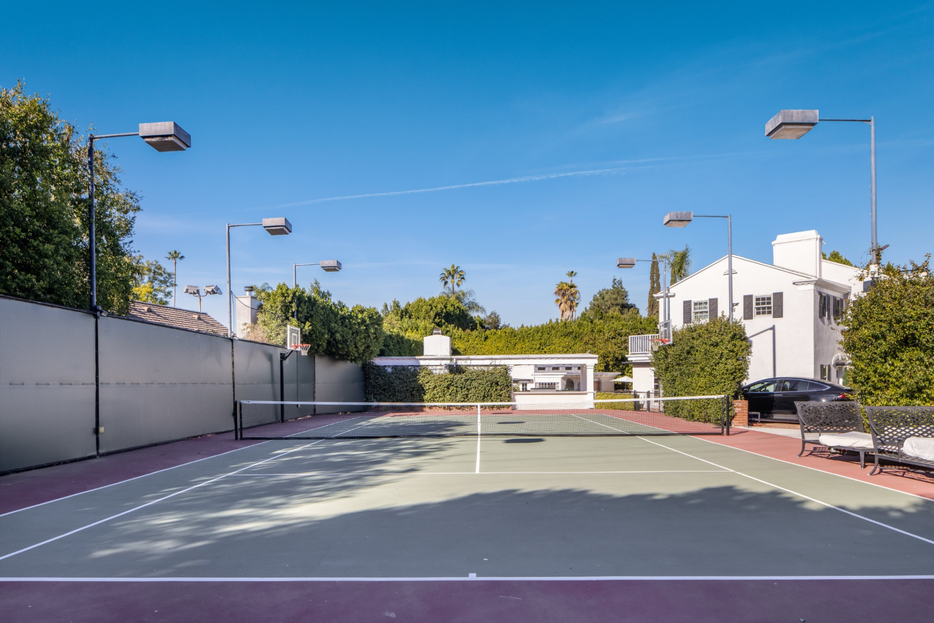 Off to the side of the house, a full-size tennis court is enclosed by tall walls and hedges, with lighting extending around its perimeter for evening play.