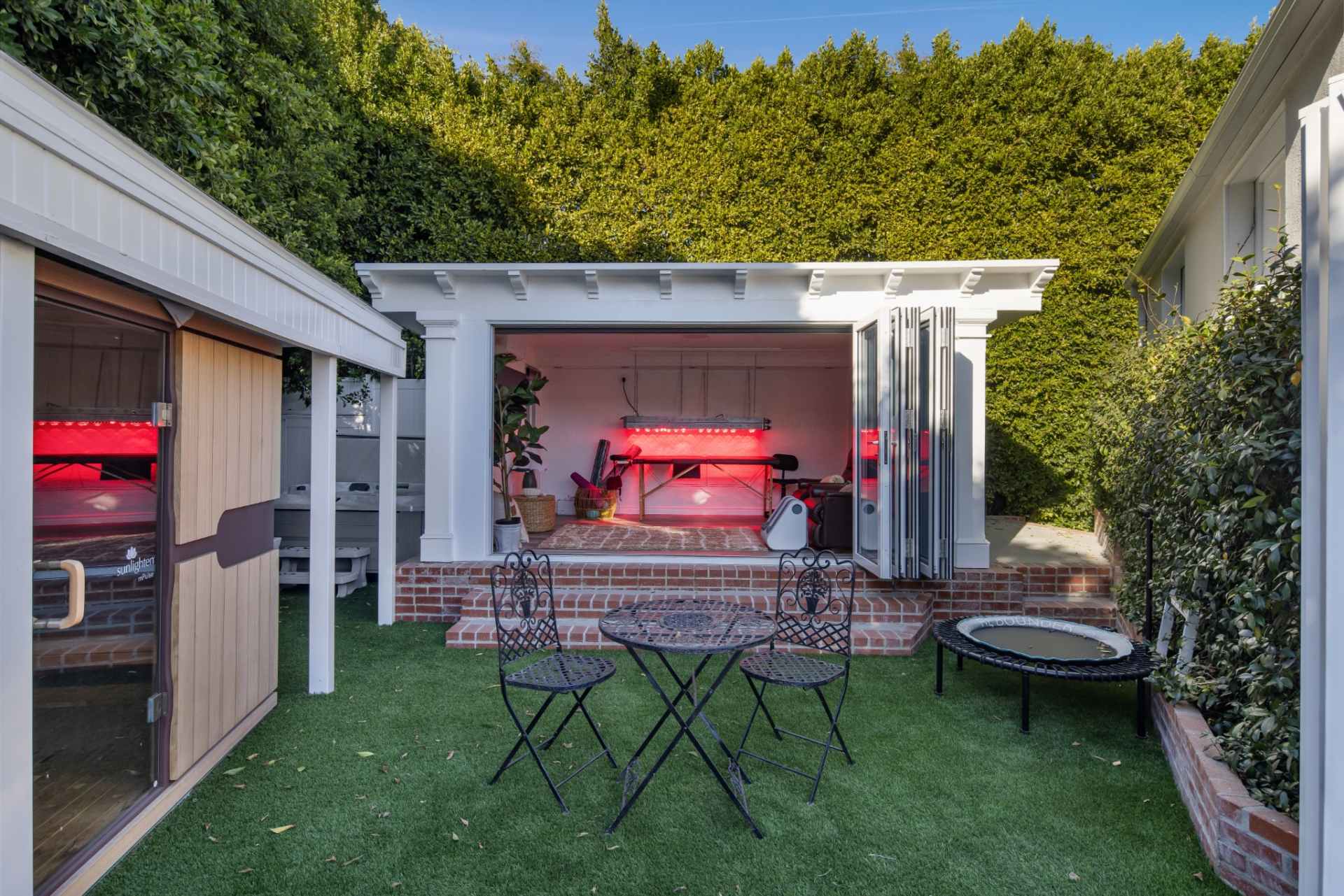 One structure opens through folding glass doors to a red-light therapy room, while a separate wood-lined sauna sits directly across the turf-covered courtyard.