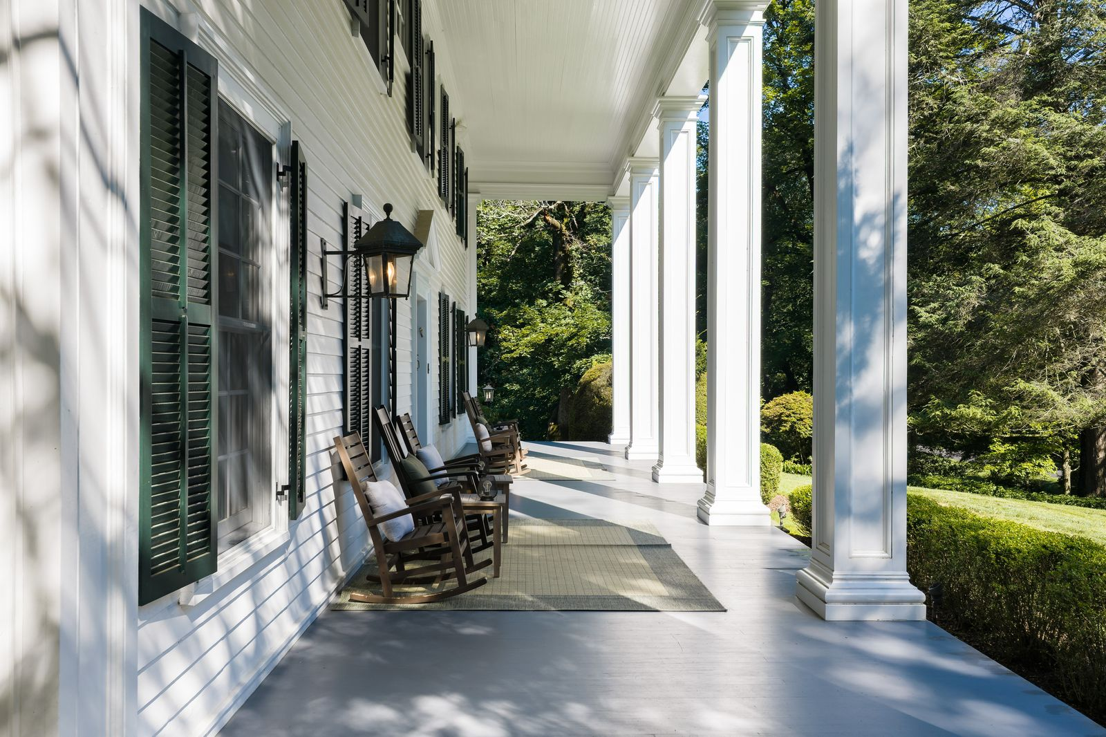 A front porch runs across the front of the house beneath a deep overhang supported by six tall columns, with black shutters, white clapboard siding, and a series of rocking chairs lining the covered walkway.