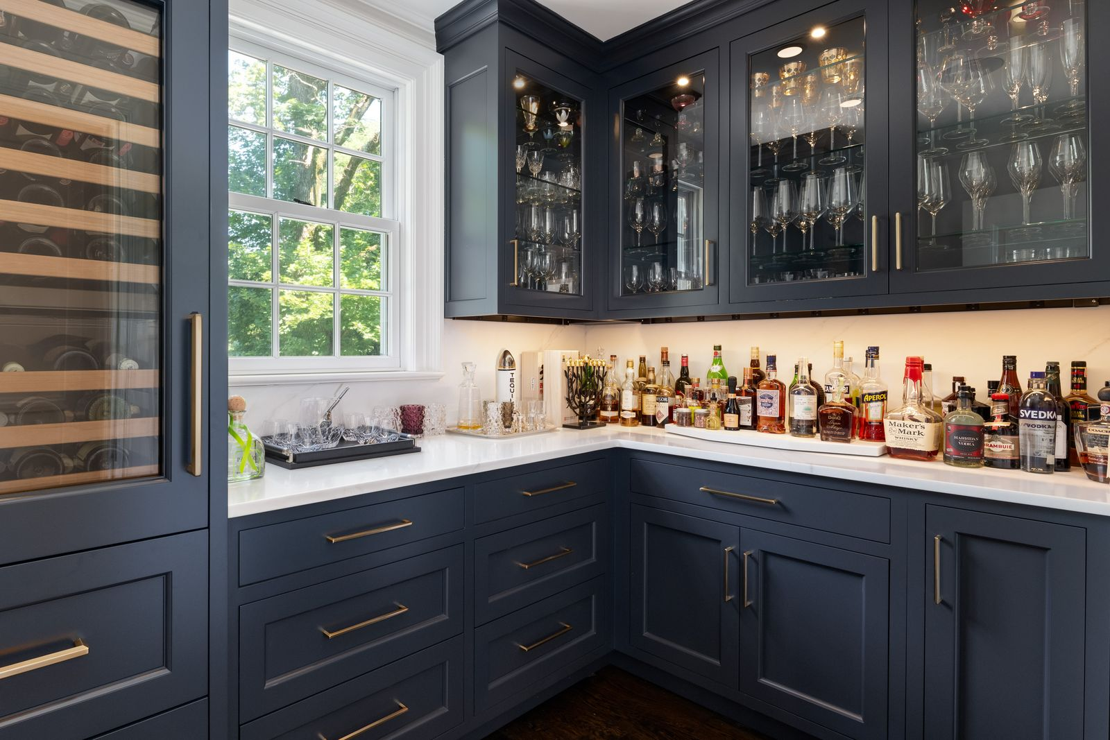 Dark blue cabinetry wraps the room on multiple sides, with glass-front upper cabinets displaying glasses above white countertops lined with drinks.