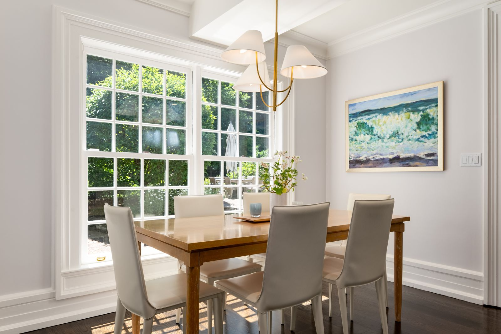 A breakfast nook, made up of a simple wood table and light upholstered chairs, sits in front of a large gridded window overlooking the garden.