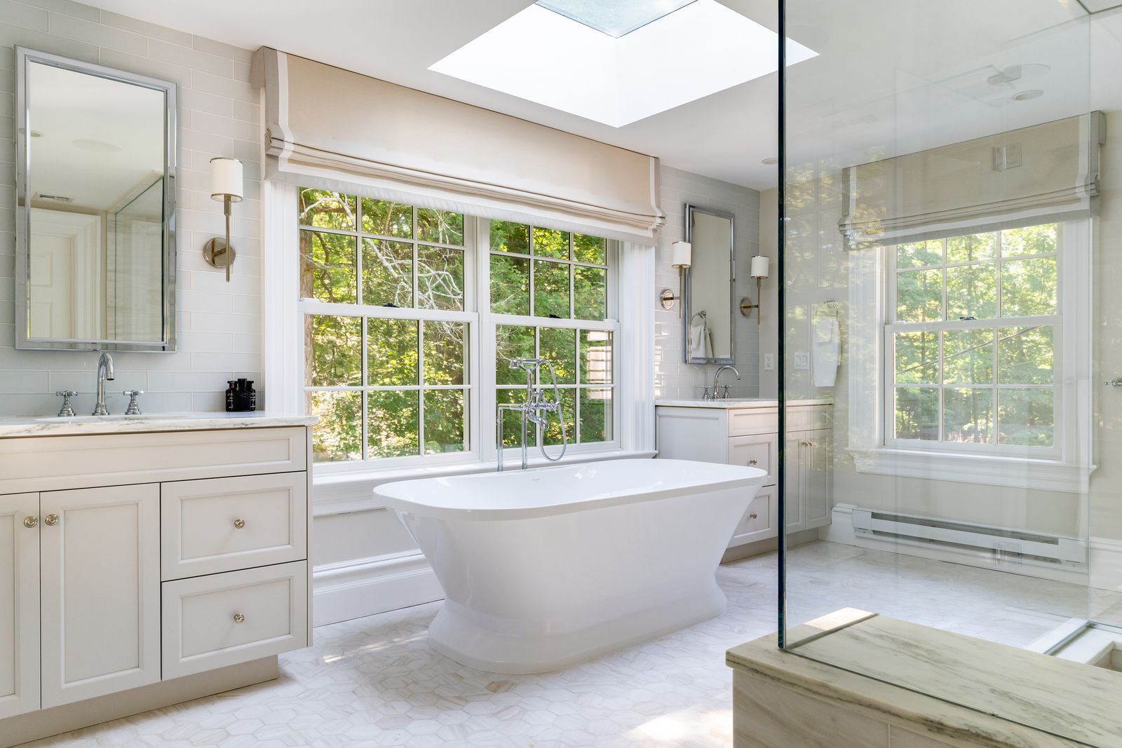 The bathroom is finished almost entirely in pale marble, with a freestanding soaking tub positioned in front of a large window and beneath a skylight. 