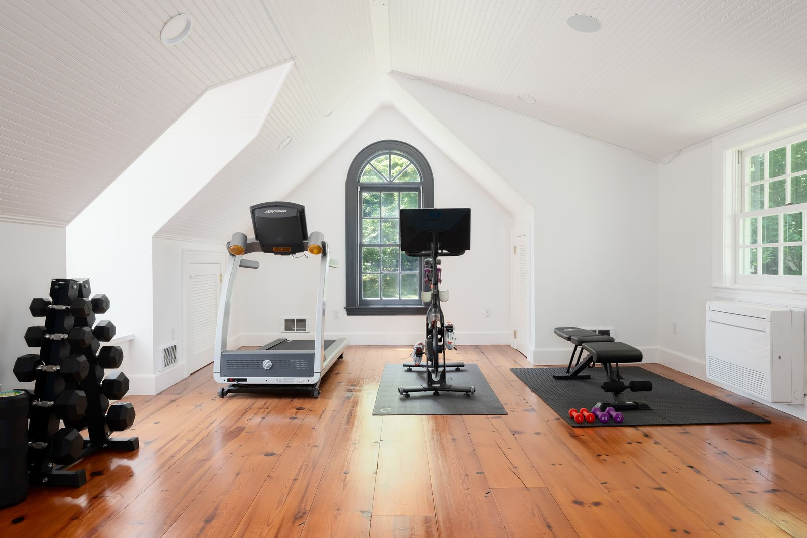 A tall arched window at one end and additional side windows bring in natural light, while the open floor plan leaves room for cardio equipment, free weights, and a workout bench.