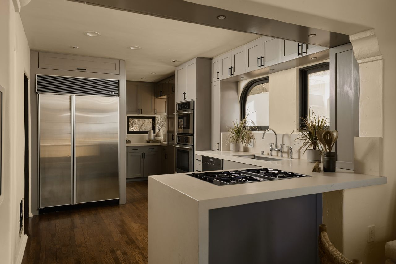 At the far end, a large stainless-steel refrigerator and stacked wall ovens are built into the cabinetry, with additional storage continuing into the back corner.