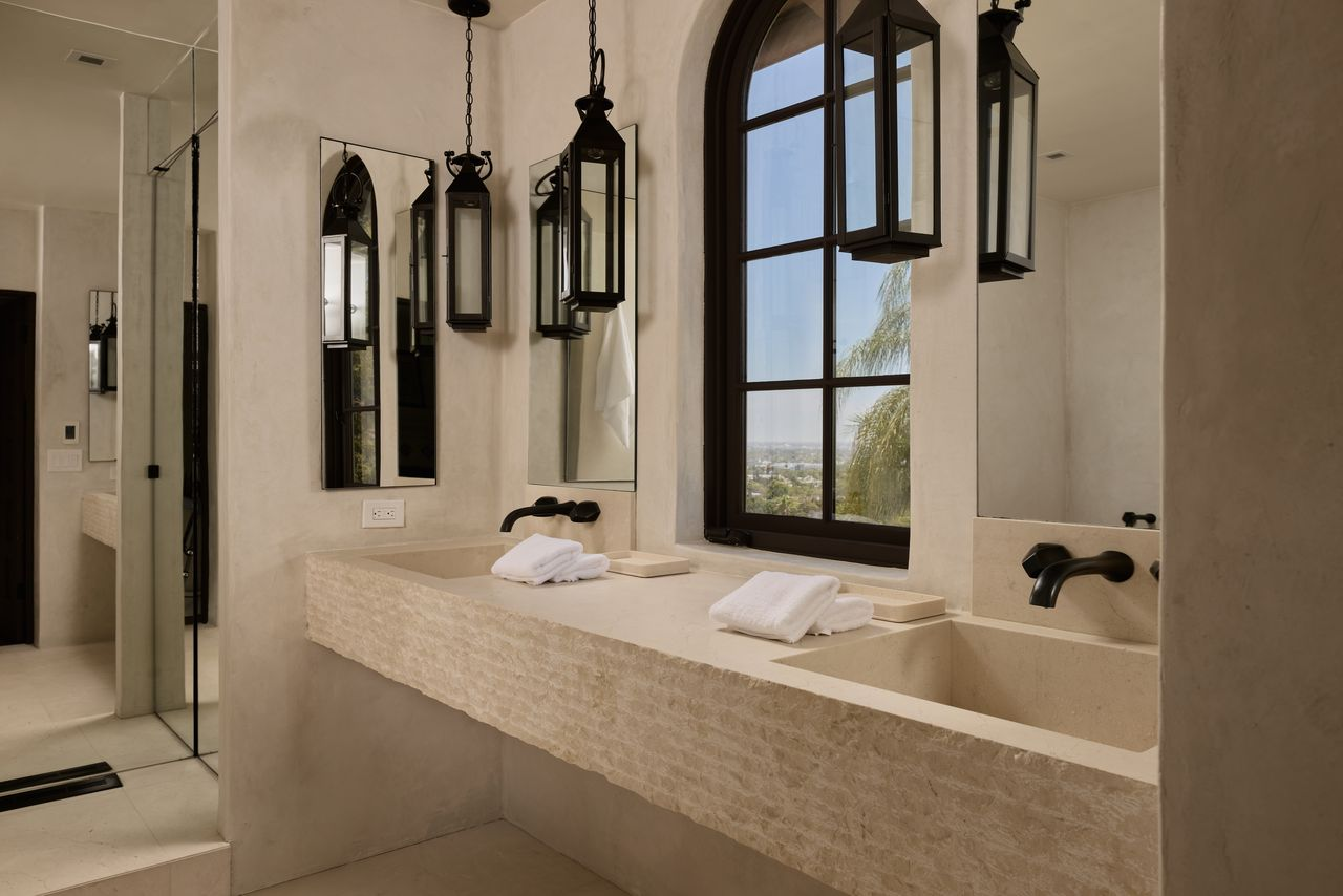 Pale stone surfaces line a long double vanity set beneath separate mirrors, an arched window, and pendant-style lanterns.