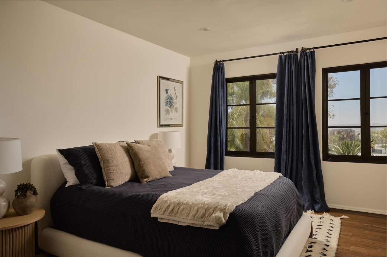 In the middle of the room, a low upholstered bed sits parallel to the windows with matching dark blue bedding and neutral accents.