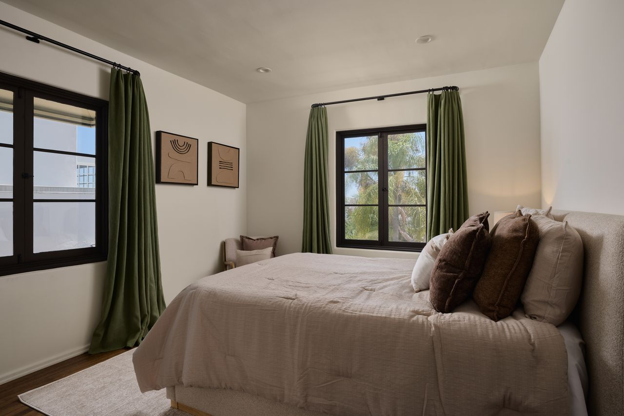 The second bedroom has a similar layout, but its windows are placed on different walls framed by green drapery, and the bed has soft pink bedding.