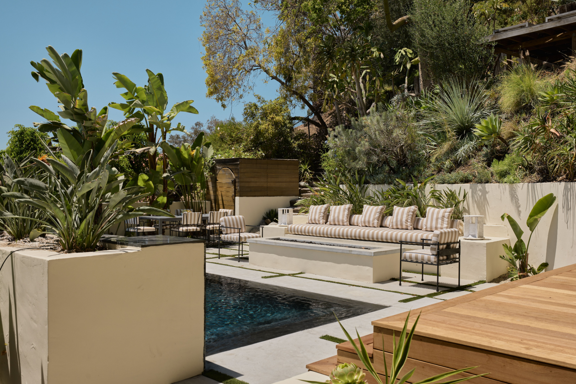 A long built-in sofa faces a rectangular fire pit, with a dining table sitting farther back near the landscaping.