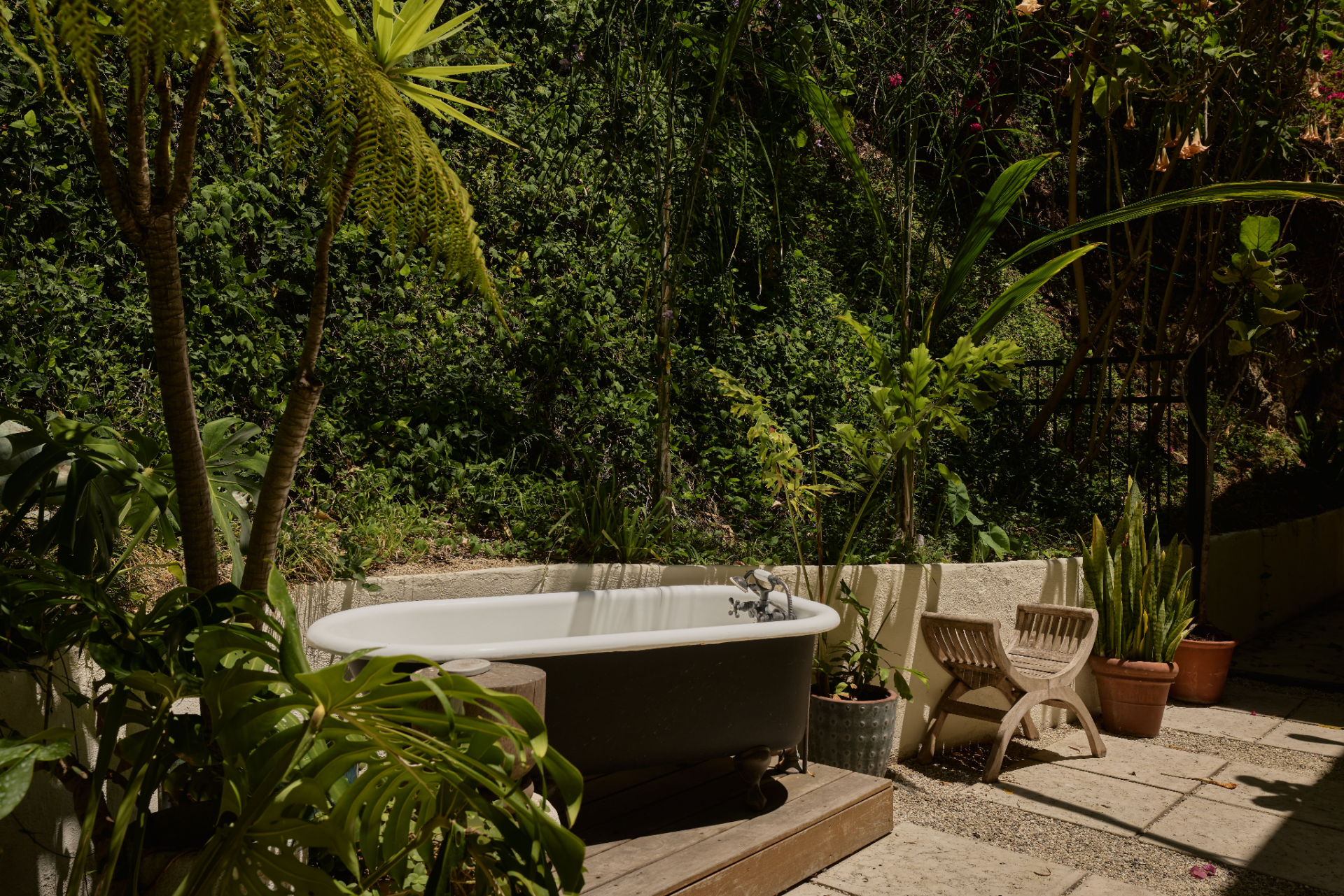 A freestanding claw-foot tub rests on a small wood platform, creating an outdoor bathing area surrounded by tropical greenery.