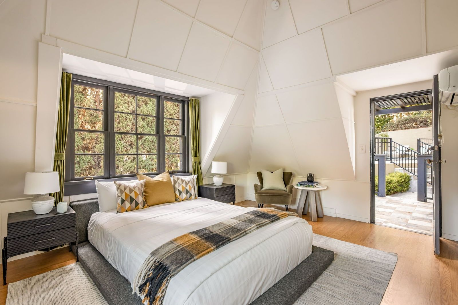 The sleeping area sits beneath a wide bank of black-framed windows, with the bed positioned between two nightstands and an additional sitting area tucked along the opposite wall.