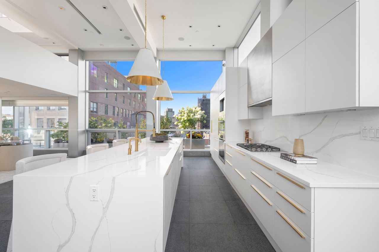 A long counter and cooktop line the back wall beneath additional cabinetry, while brass hardware and fixtures add a warmer contrast to the white palette.