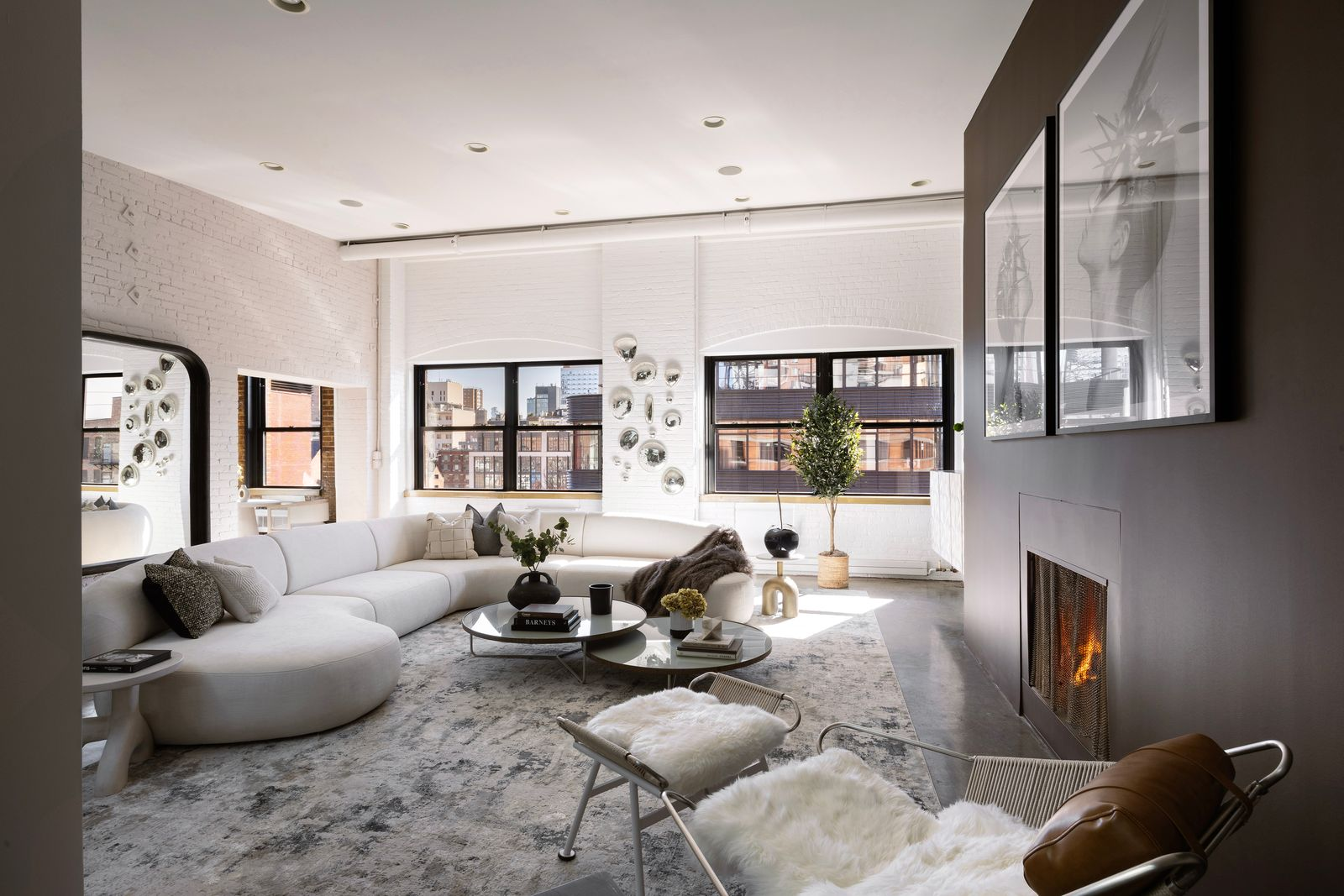 The living area gathers around a wood-burning fireplace, while white exposed brick and large black-framed windows line the surrounding walls.