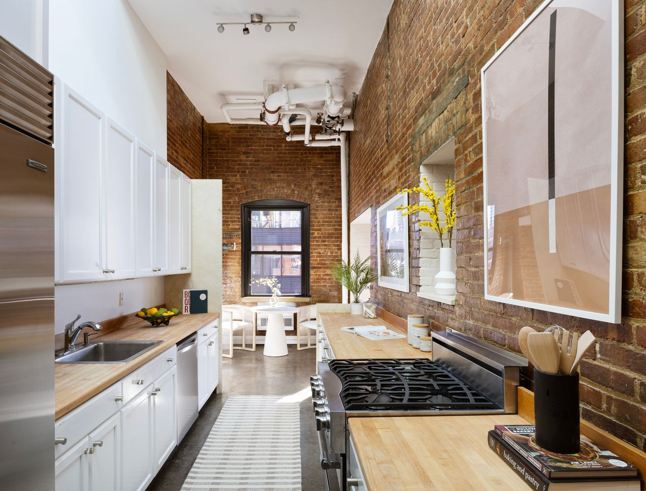 The kitchen narrows into a galley-style space where exposed brick runs along one wall opposite white cabinetry topped with butcher-block counters. 