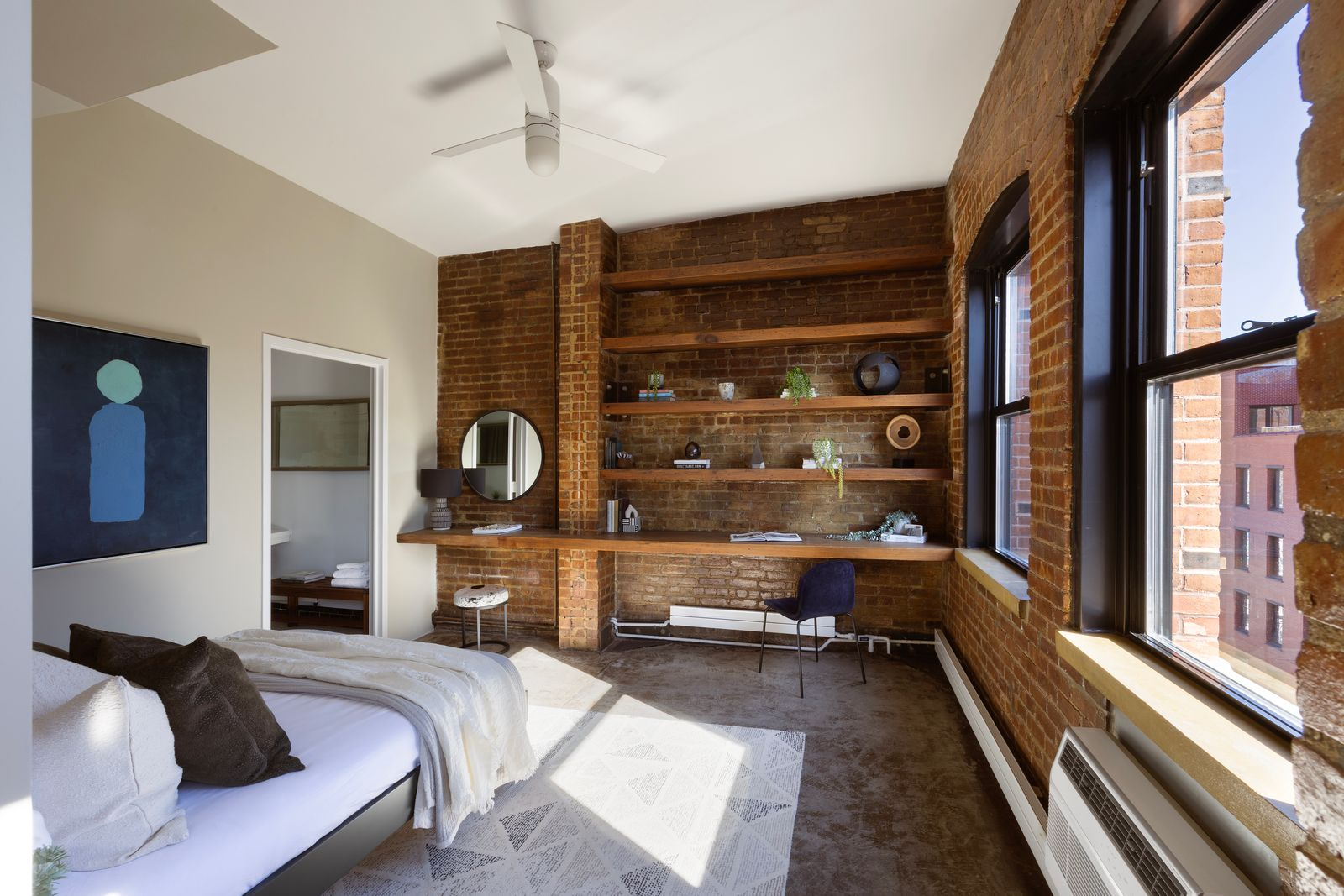 The guest suite keeps the exposed brick intact along two walls, where built-in wood shelving and a long work surface create a desk and vanity area. 