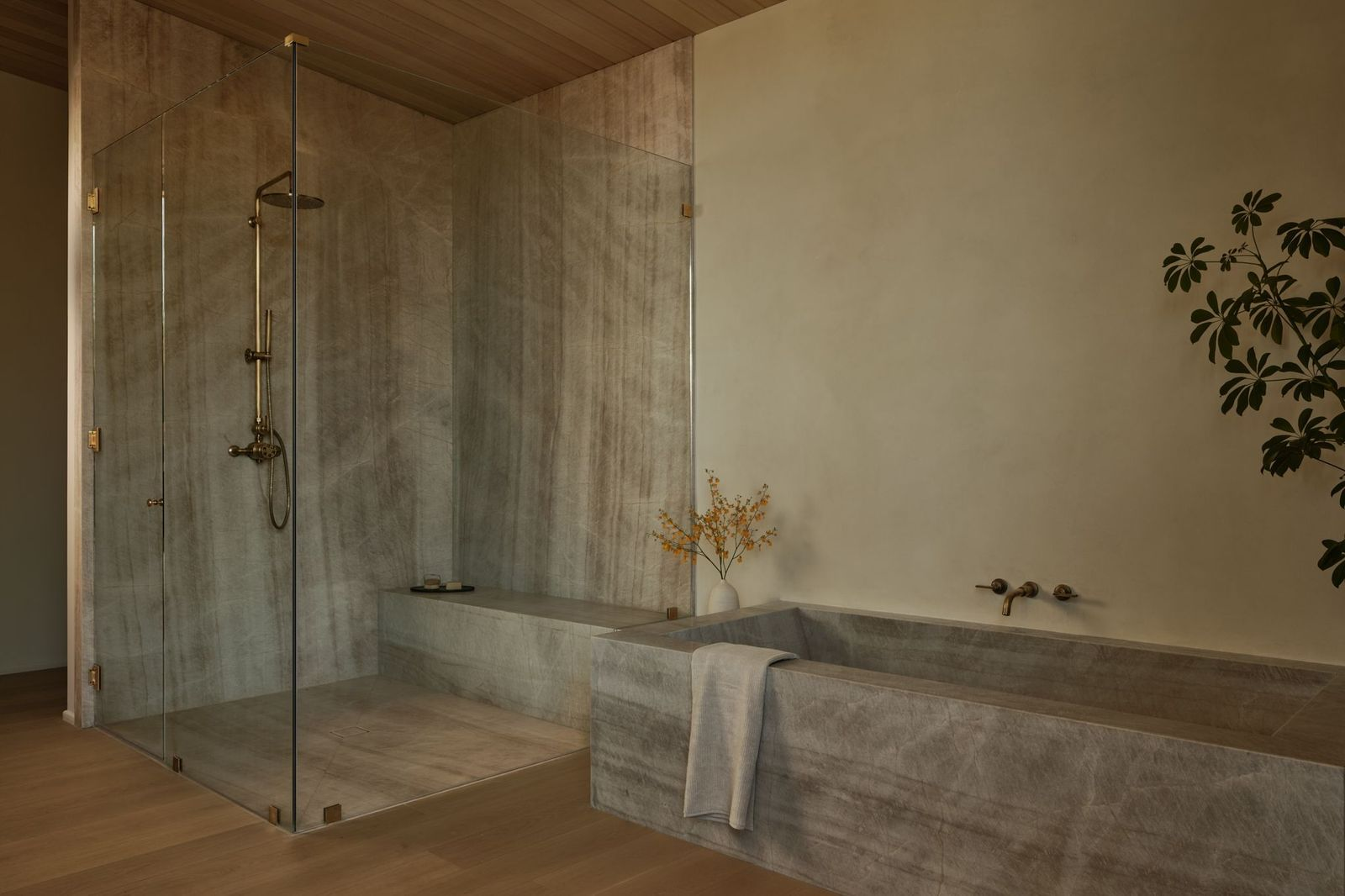 A stone soaking tub runs beside a glass-enclosed shower, with the same grey stone extending across the shower walls, bench, and tub.