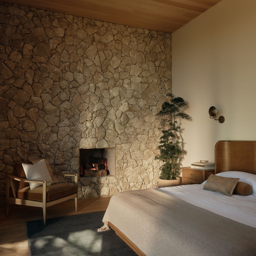 The bed sits next to a full wall of stone surrounding a wood-burning fireplace.