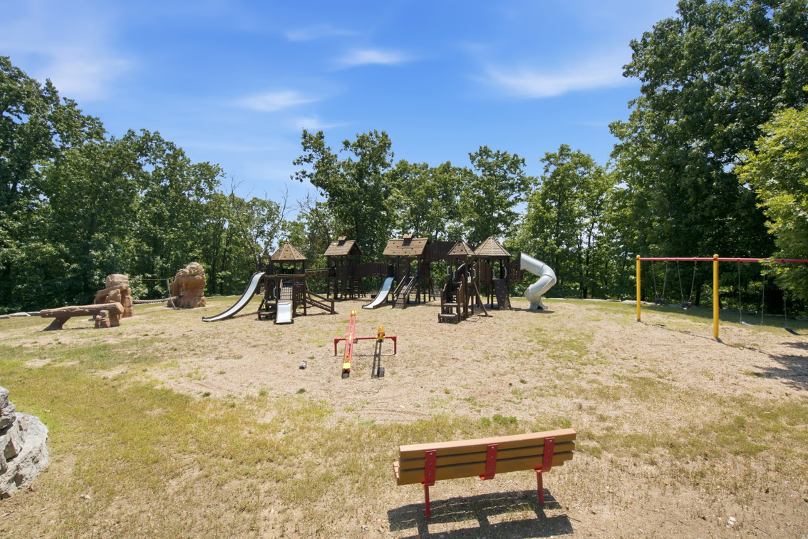 Multiple slides, climbing structures, swings, seesaws, and additional play features spread across the open lawn, with tall trees surrounding the area.
