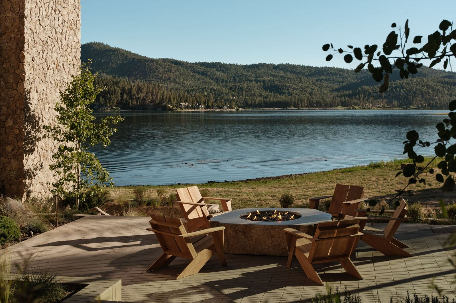 Wooden chairs circle the stone fire pit with the lawn and lake directly past it.