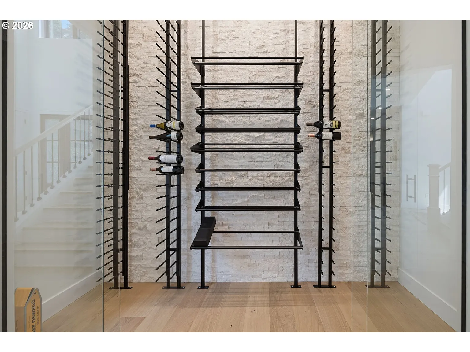 A glass-enclosed wine gallery with black metal bottle racks rising nearly the full height of the room against a textured stone wall.