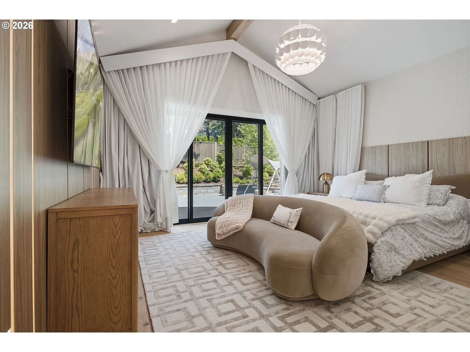 The bed and a curved couch at its foot face a mounted TV while layered drapery frames black-framed glass doors that open to the backyard. 