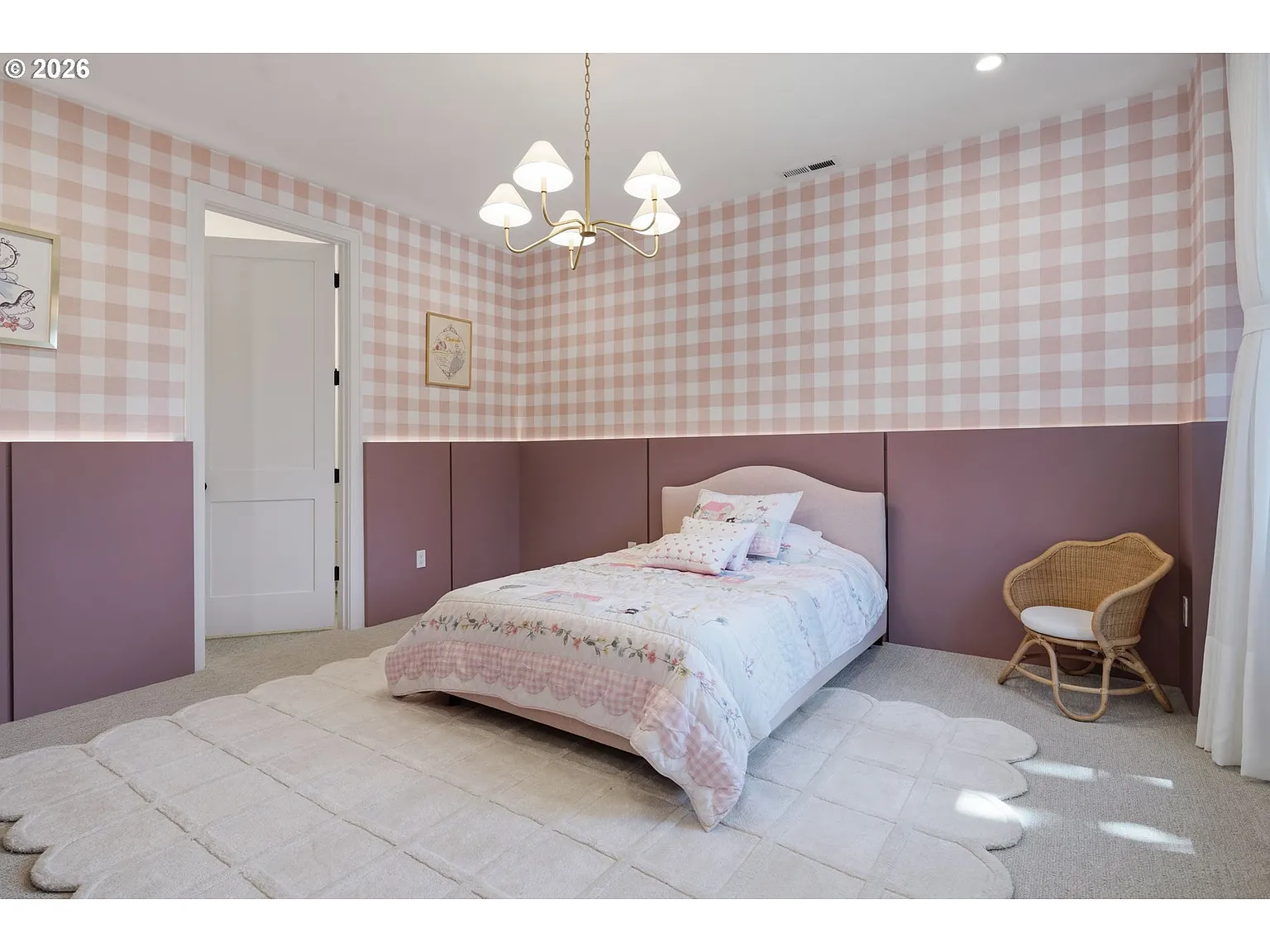 The first bedroom has a playful, tailored look with soft pink plaid wallpaper wrapping the room above a deeper rose-colored paneled wainscot. 
