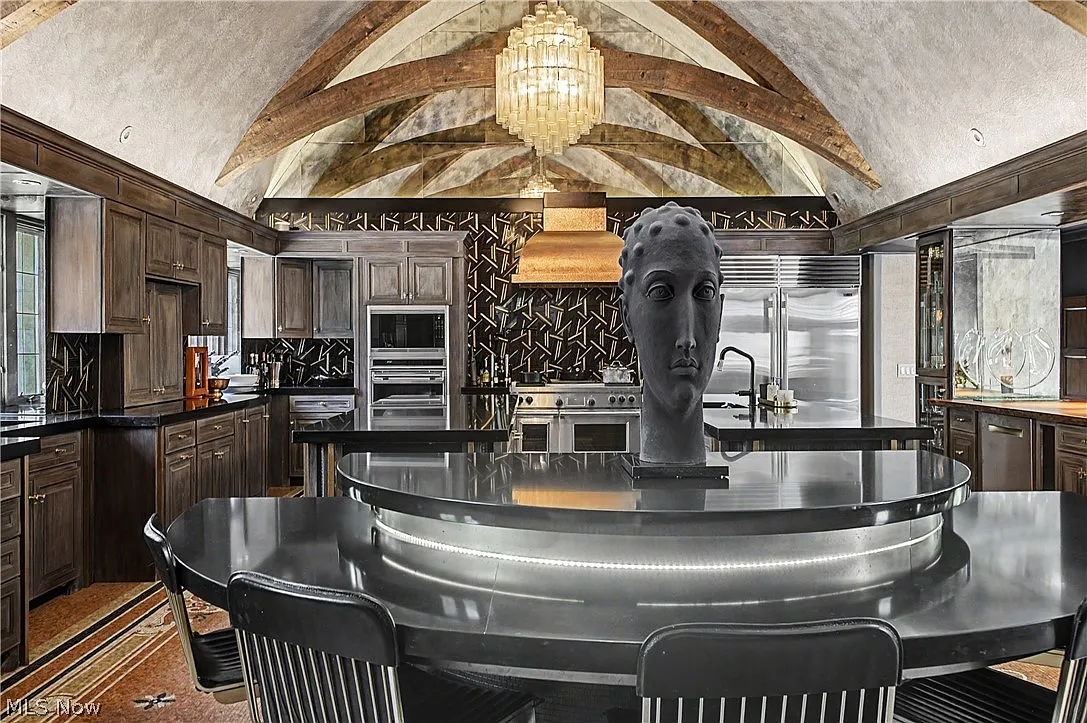 Dark wood cabinetry wraps the perimeter beneath a vaulted ceiling crossed by exposed timber beams, with the room centered around two large stainless-steel islands and a curved breakfast nook.