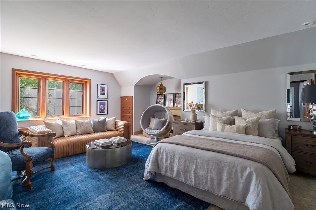Inside the bedroom, a bed is set to one side of the room, while a leather sofa, lounge chair, and circular chair create a separate sitting area near the windows.