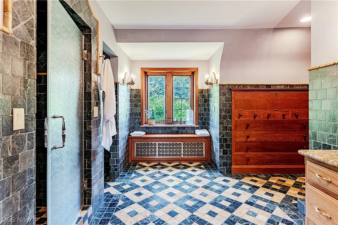 A large window at the far end brings natural light into the room above a built-in bench, while the layout extends toward a glass-enclosed shower and additional vanity space.