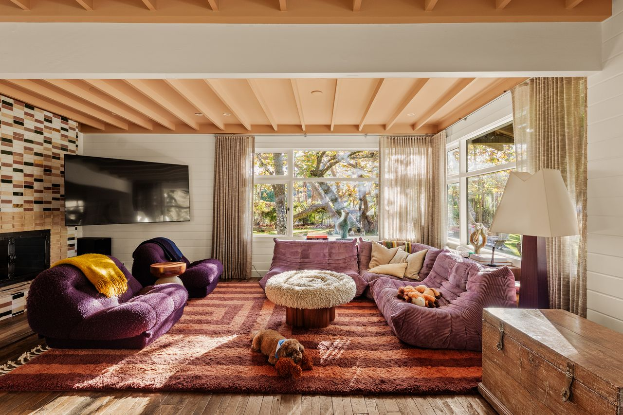 Within the space, a curved purple couch is tucked beneath the windows, and a pair of darker purple lounge chairs is placed in front of the fireplace.