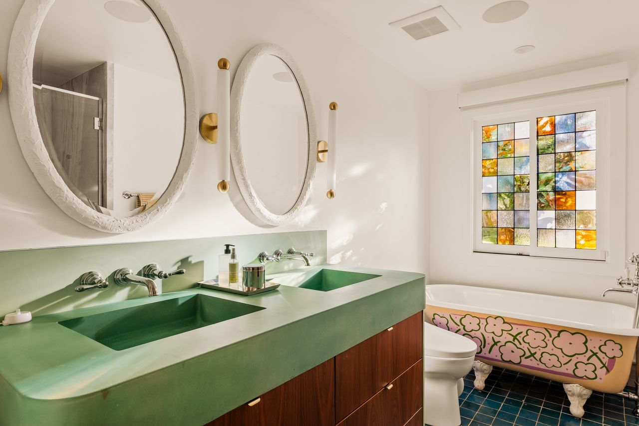 On the back wall, a clawfoot soaking tub sits beneath a stained-glass window, while the separate marble shower sits across from it.