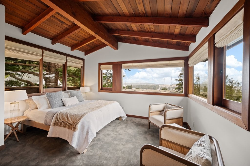 Exposed wood beams continue across the ceiling, while the bed is positioned along the window-lined corner.