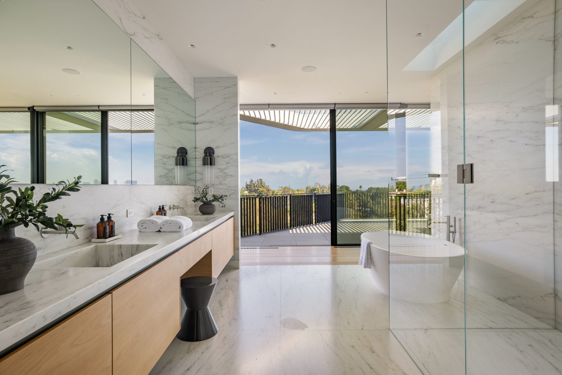 Bathroom is finished in pale stone with a long double vanity, freestanding soaking tub, and glass-enclosed shower.