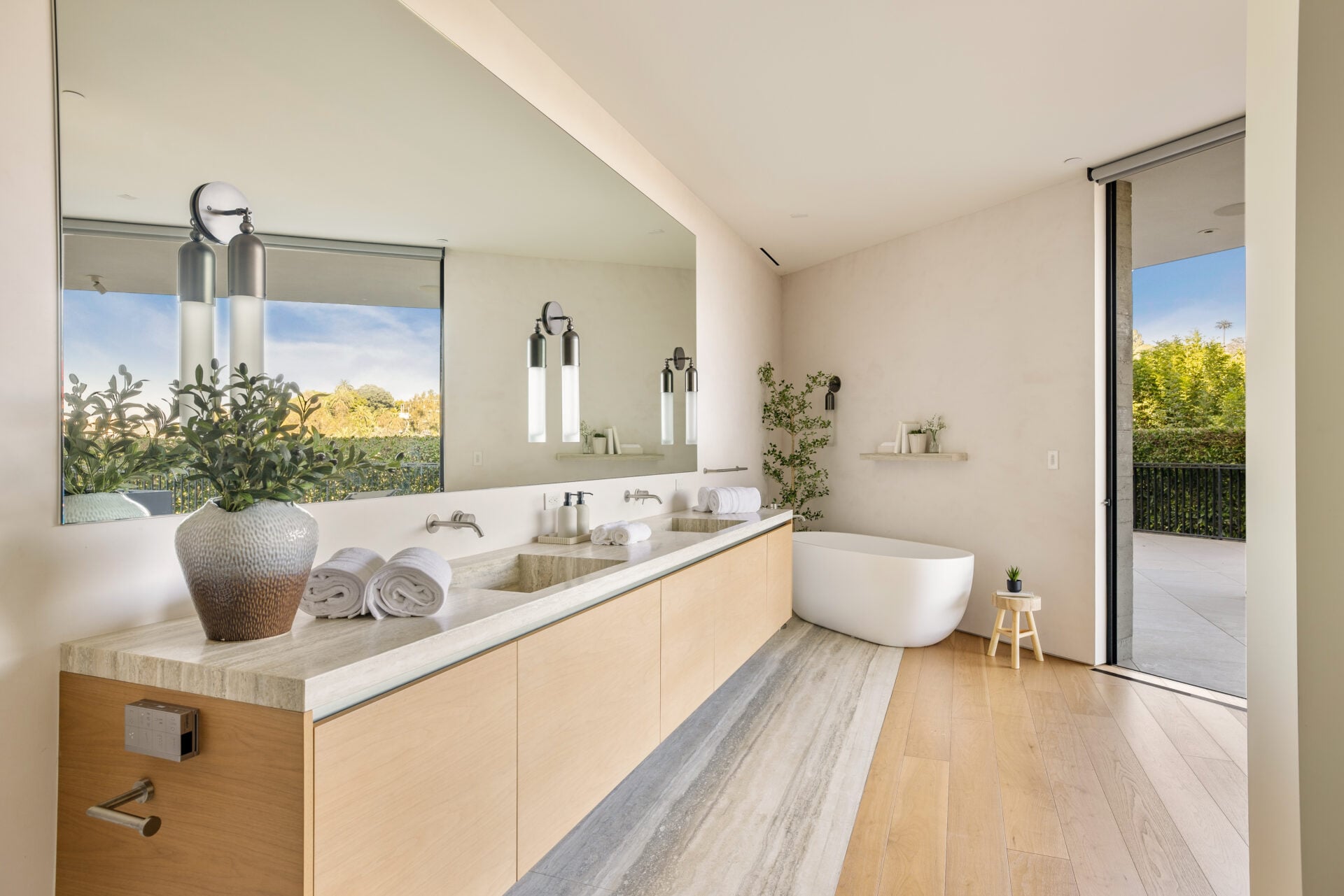This suite's bathroom has a warmer palette, pairing a long wood-fronted double vanity with a freestanding tub positioned near its own exterior opening.